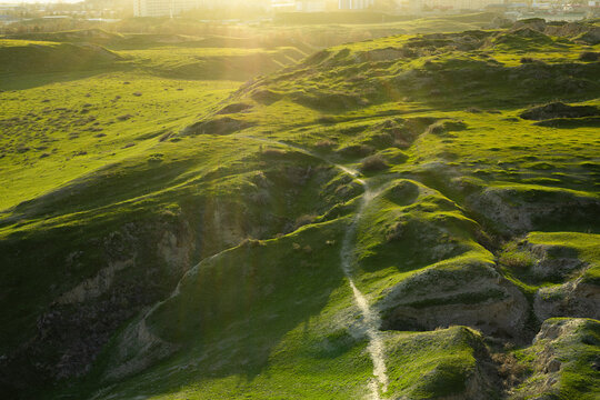 Green hills of Afrasiyab archaeological site near Samarkand at golden hour. Ancient eroded landscape with footpath contrasts with modern city skyline, ideal for travel and heritage publications.