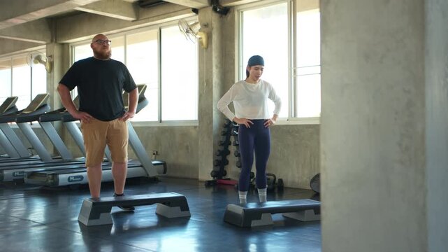 Personal trainer and overweight beginner performing step aerobics exercises together in a fitness gym