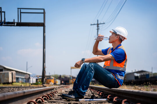 Construction worker drinks water on railroad maintenance site, Railway worker sitting on tracks drinking water to stay hydrated under hot sun, Tired technician taking a break at railway depot