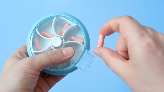 Female hands holding a pill case with pink capsules close-up on a blue background.