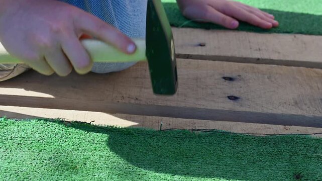 a person's hands using a hammer to carefully drive a small nail into a rough wooden plank, securing a piece of bright green artificial turf.