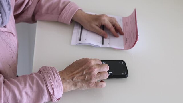 Senior woman using a smartphone calculator while checking household bills at home. Close-up of monthly expenses, personal budgeting and cost of living.