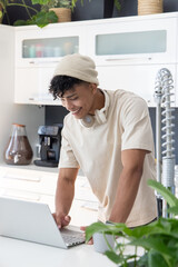 African American man in 20s leaning over laptop in kitchen, smiling with beanie and headphones