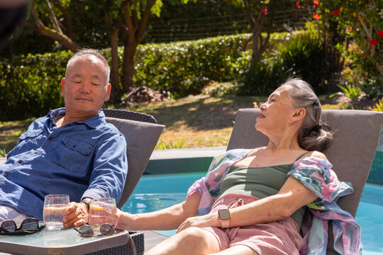 Senior Asian couple reclining poolside holding hands on table with lemonade, in shirt and swimsuit