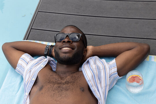 African American man reclining poolside on chair, in striped shirt and shades, holding citrus glass