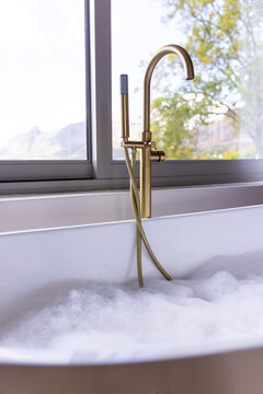 Brushed-gold bathtub faucet with hand sprayer is resting on foamy tub rim by window, mountain view