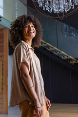 Obraz premium African American woman standing, smiling and looking upward in atrium with chandelier and bracelets
