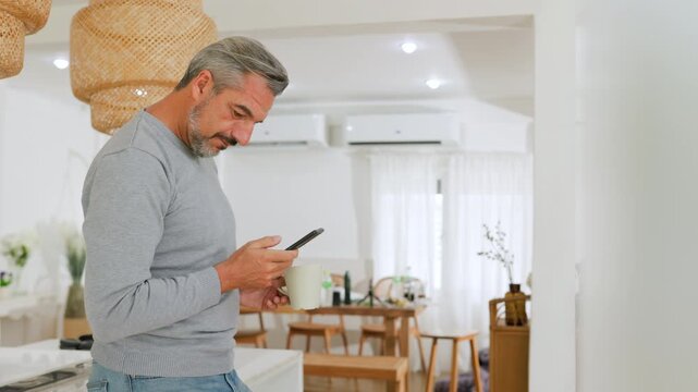 Mature man checks a smartphone while holding a cup of coffee in a bright modern home, reflecting connected daily living.