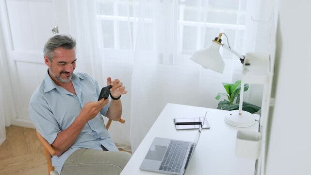 Smiling businessman uses a smartphone in a bright home office with a laptop, reflecting flexible remote work and digital communication.