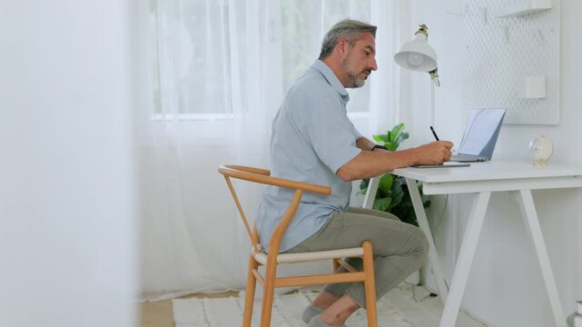 Mature businessman works at a laptop while taking notes in a bright home office, highlighting productivity and remote work.