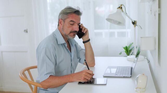 Mature businessman works from a home office, talking on a smartphone while taking notes beside a laptop.