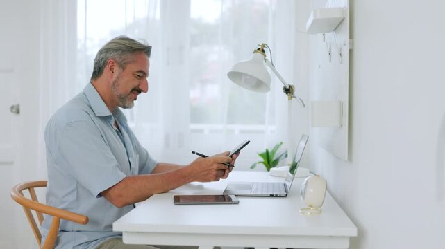 Mature businessman records a voice message while taking notes beside a laptop in a home office, showing remote work and digital communication.