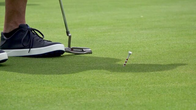 A close-up shot captures a golfer using a putter to hit a white ball slowly across the green grass. A red and white striped tee stands stuck in the turf.