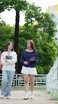 Two female university students walk across campus carrying tablets while talking, representing teamwork and modern education.