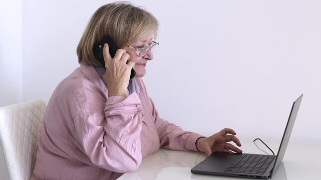 Smiling senior woman talking on smartphone while using a laptop at home. Mature woman getting online support, working remotely or communicating with family.