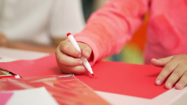 Child uses a red marker to draw on bright orange paper during a creative classroom activity, highlighting learning and fine motor skills