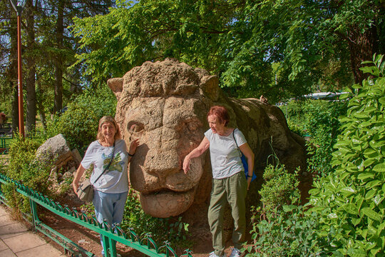 Two female tourists smile happily, posing next to the famous stone sculpture of the Lion of Ifrane in Morocco.