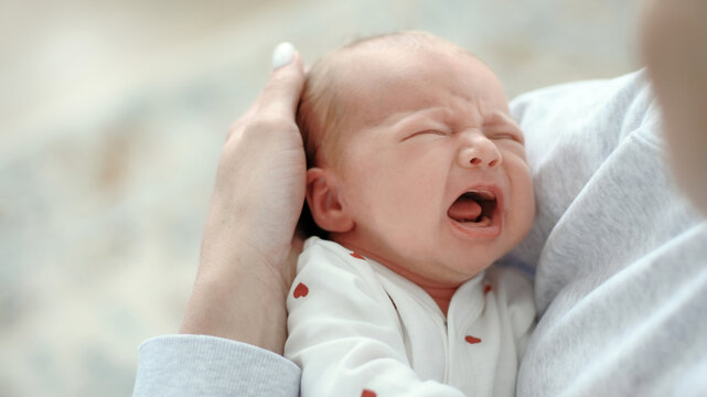 Infant cries loudly, mouth open wide, eyes shut tight. Mom comforts baby, supporting head with gentle hand indoors.