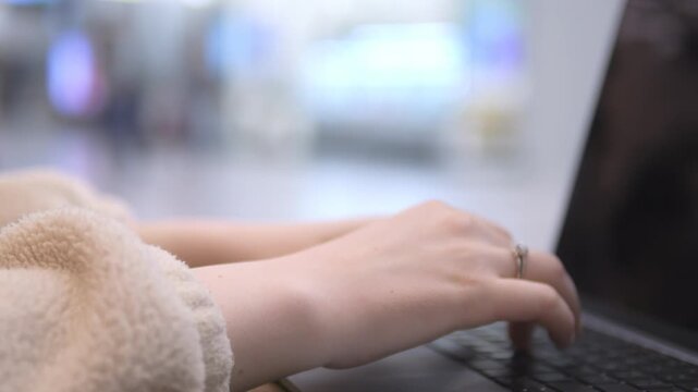 Close-up of hands typing on a laptop. Person wears soft beige sleeve. Background softly blurred. Ring visible on finger. Focus remains on hands and keyboard