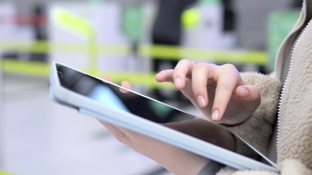 Person holds tablet, fingers tap screen. Light jacket, blurred background with yellow barriers. Neutral indoor lighting, focus on hands and device