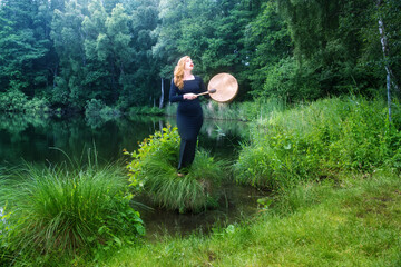 Young redhead woman playing a shamanic frame drum beside a tranquil forest lake, creating a spiritual atmosphere of meditation, rhythm, nature and inner connection © ullision