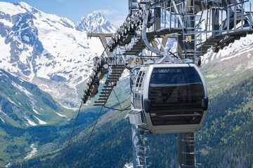 Mountain cable car gondola travels above a green valley with trees, showcasing snow-covered mountains in the distance and a bright blue sky © Anat art