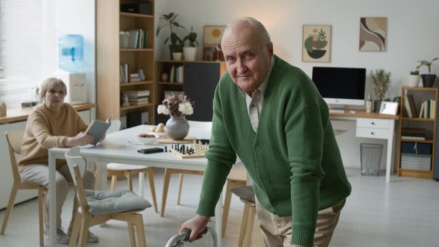 Portrait of senior man using walker in retirement home and looking at camera, senior woman reads in background. Footage for senior care and healthcare marketing