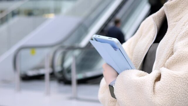 Person holds tablet while standing near escalator. Background shows moving escalator with blurred people. Focus on tablet and person's coat. Modern indoor setting with neutral lighting