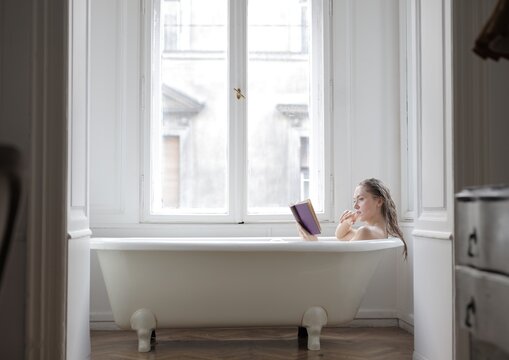 Young woman reading a book in a bathtub by a large window