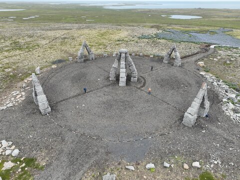 Aerial view of Arctic Henge, a modern stone monument with towering arches arranged in a circle on a gravel landscape, with people walking nearby in Raufarhofn, Nordurthing, Iceland.