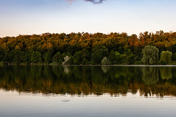 Still lake reflects a belt of summer forest under a pale evening sky with gentle ripples adding texture to the glassy surface creating a peaceful nature scene perfect for calm outdoor themes © Chornogirskyi
