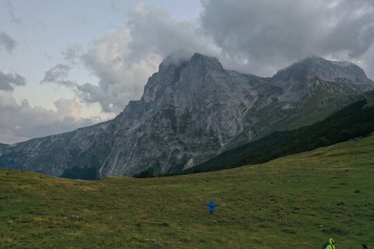 Aerial view of the Gran Sasso massif at Prati di Tivo, featuring rugged mountain peaks, clouds, green slopes, and a person standing in the foreground. Prati di Tivo, Abruzzo, Italy.