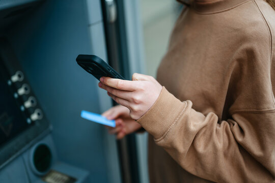 Woman using smartphone and credit card at atm for cash withdrawal