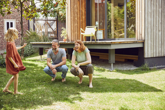 Cheerful parents looking at daughter while crouching on grassy land in yard