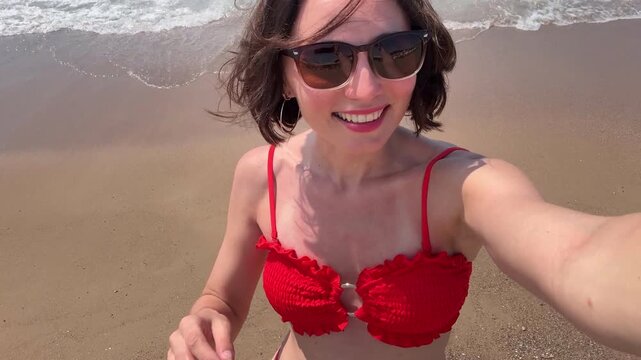 Close up portrait of a smiling middle aged caucasian woman in red textured bikini and sunglasses taking a selfie on a sandy coast with sea water wave