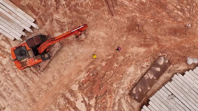 Construction Workers Walking Between Excavator and Foundation Piles. A bright orange excavator and rows of concrete piles frame a group of workers crossing an active construction site.