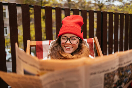 Smiling woman wearing knit hat and eyeglasses reading newspaper while sitting at sidewalk cafe