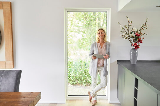Portrait of happy woman standing at French door using tablet at home