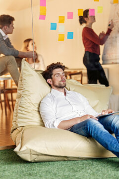 Relaxed man in office sitting in bean bag with meeting in background