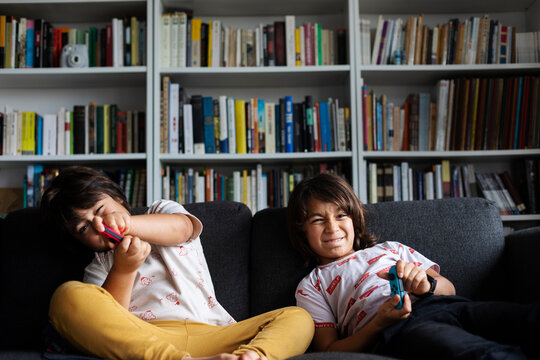 Siblings playing video game while sitting on sofa against bookshelf in living room at home