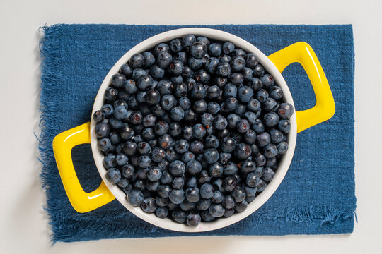 Fresh blueberries in bowl, close up. Top view of round bowl filled with ripe wild blueberries on blue fabric napkin over white background representing healthy eating, summer harvest and organic food