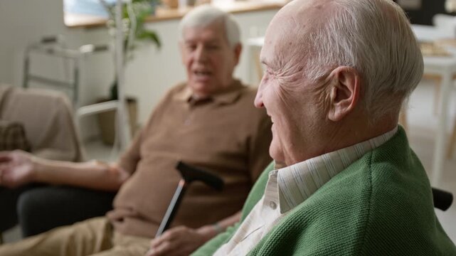 Side view shot of two senior men sitting in retirement home, talking and laughing together during social activity. For promoting assisted living facilities, geriatric healthcare