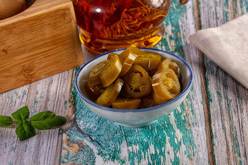 Fototapeta premium Sliced pickled jalapeno peppers in a bowl on a rustic wooden table with chili oil and fresh basil
