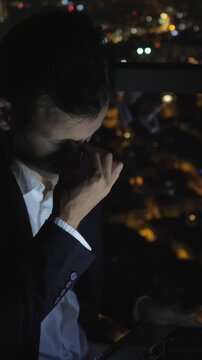 Tired businessman working on a tablet late into the night. Feeling stressed and exhausted while overlooking the illuminated cityscape from a high rise office. Eventually rubbing his eyes and yawning