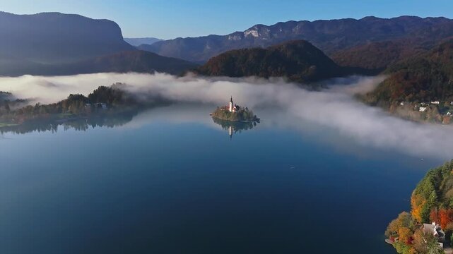 Bled, Slovenia - 4K Flying towards the famous Bled Island on Bled Lake on a sunny, foggy autumn morning with Julian Alps and blue sky at background
