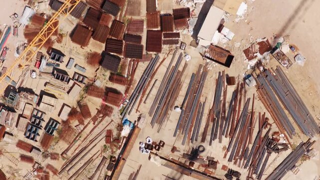 High-angle aerial perspective flying over a construction site supply yard, showing stacks of rusty rebar, metal beams, and the shadow of a large crane moving across the dusty ground