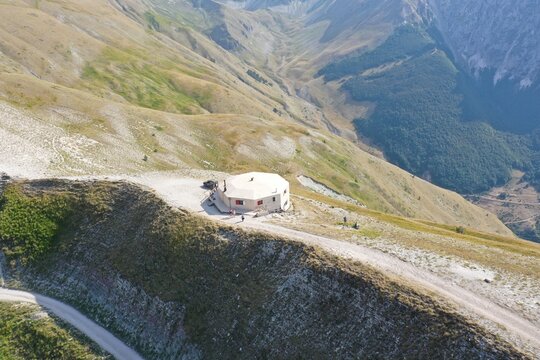 Aerial view of the mountain refuge along the Strada per Rifugio del Fargno, surrounded by the rugged slopes and grassy terrain of the Sibillini Mountains in Ussita, Marche, Italy.