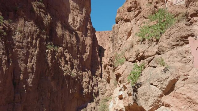 In Morocco's Todra Gorge near Tinghir, towering vertical cliffs and red rock formations rise above the arid desert canyon and rocky terrain under a clear blue sky.