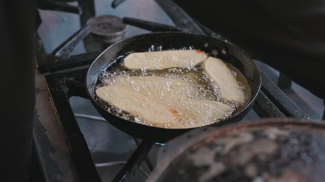 Chef wearing protective gloves fries potato wedges in hot oil as bubbles rise across the pan during commercial food preparation, close-up cooking shot.