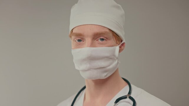 Caucasian doctor wearing surgical mask closeup, stethoscope visible, protective face covering in clinical setting, frontline